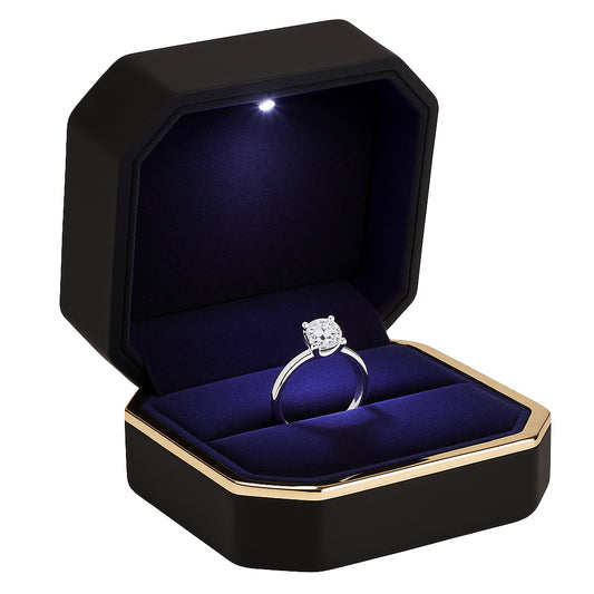 Open black matte ring box with gold trim and LED light holding diamond engagement ring